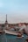 A panoramic view of Parisian rooftops with the Eiffel Tower in the background. The rooftops are characterized by their blue and green hues and visible chimneys. Smoke rises from one of the chimneys, and the sky is overcast with a hint of blue peeking through.