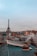 A panoramic view of Parisian rooftops with the Eiffel Tower in the background. The rooftops are characterized by their blue and green hues and visible chimneys. Smoke rises from one of the chimneys, and the sky is overcast with a hint of blue peeking through.