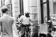 A black and white street scene featuring a police officer on a bicycle interacting with a man sitting on the sidewalk holding a cardboard sign that reads 'HOMELESS ANYTHING HELPS Please Thank You'. Other pedestrians are visible walking past in the background, and the setting appears to be urban with storefronts and a window display.