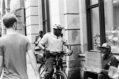 A black and white street scene featuring a police officer on a bicycle interacting with a man sitting on the sidewalk holding a cardboard sign that reads 'HOMELESS ANYTHING HELPS Please Thank You'. Other pedestrians are visible walking past in the background, and the setting appears to be urban with storefronts and a window display.