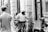 A black and white street scene featuring a police officer on a bicycle interacting with a man sitting on the sidewalk holding a cardboard sign that reads 'HOMELESS ANYTHING HELPS Please Thank You'. Other pedestrians are visible walking past in the background, and the setting appears to be urban with storefronts and a window display.