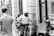 A black and white street scene featuring a police officer on a bicycle interacting with a man sitting on the sidewalk holding a cardboard sign that reads 'HOMELESS ANYTHING HELPS Please Thank You'. Other pedestrians are visible walking past in the background, and the setting appears to be urban with storefronts and a window display.