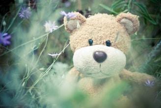 A plush teddy bear is nestled among wildflowers and tall grasses, with soft sunlight filtering through the greenery. The bear's fur is light brown, and it has a rounded nose and black eyes, exuding a sense of comfort and nostalgia. The background includes hints of purple flowers and lush greenery, creating a serene and natural setting.