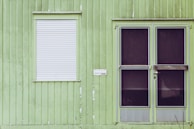 The image depicts a green exterior wall of a building with vertical panel siding. There is a white roll-down shutter covering the window on the left, and a double door with dark, almost opaque, glass panels on the right. There is also a small white mailbox or nameplate mounted on the wall between the window and the door.