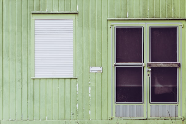 The image depicts a green exterior wall of a building with vertical panel siding. There is a white roll-down shutter covering the window on the left, and a double door with dark, almost opaque, glass panels on the right. There is also a small white mailbox or nameplate mounted on the wall between the window and the door.