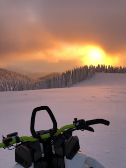 A snow-covered landscape with a snowmobile in the foreground. The scene features a dramatic sunset, casting warm orange and yellow hues over the snowy terrain. A line of frosted trees stretches along the horizon, adding to the serene winter atmosphere.