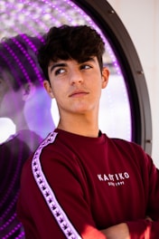 A young person with short, dark hair is wearing a maroon sweatshirt with white text and patterned stripes on the sleeves. They are standing in front of a reflective surface with purple LED lights, creating an interesting visual effect. The person appears to be pensive or thoughtful.