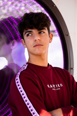 A young person with short, dark hair is wearing a maroon sweatshirt with white text and patterned stripes on the sleeves. They are standing in front of a reflective surface with purple LED lights, creating an interesting visual effect. The person appears to be pensive or thoughtful.