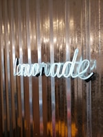 A neon sign spelling 'lemonade' is mounted on a vertically ridged metallic background. The light from the sign softly illuminates the textured surface, creating a contrast between the bright text and the darker reflective metal.
