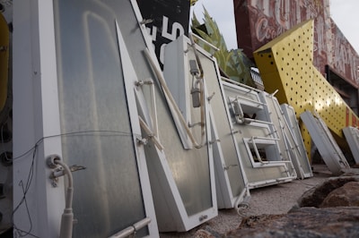 A collection of large, vintage metal letters and signs is displayed, with some lying on the ground. The signs have a weathered, retro appearance, with rust and wear visible. Some signs feature neon tubing and lights, with various colors and designs. Large, dismantled letters are stacked and lean against each other.