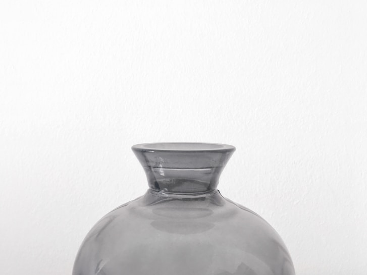 A minimalist glass vase with a slightly flared lip, made of clear, translucent material. It sits against a plain white background, emphasizing its simple and elegant design.