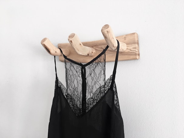 A wooden wall-mounted coat rack with three branch-shaped hooks is seen holding a black lace garment. The garment appears to be a delicate, semi-transparent piece, possibly a nightgown or slip, hanging against a plain white wall.