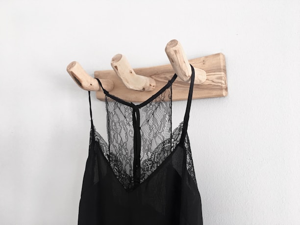 A wooden wall-mounted coat rack with three branch-shaped hooks is seen holding a black lace garment. The garment appears to be a delicate, semi-transparent piece, possibly a nightgown or slip, hanging against a plain white wall.