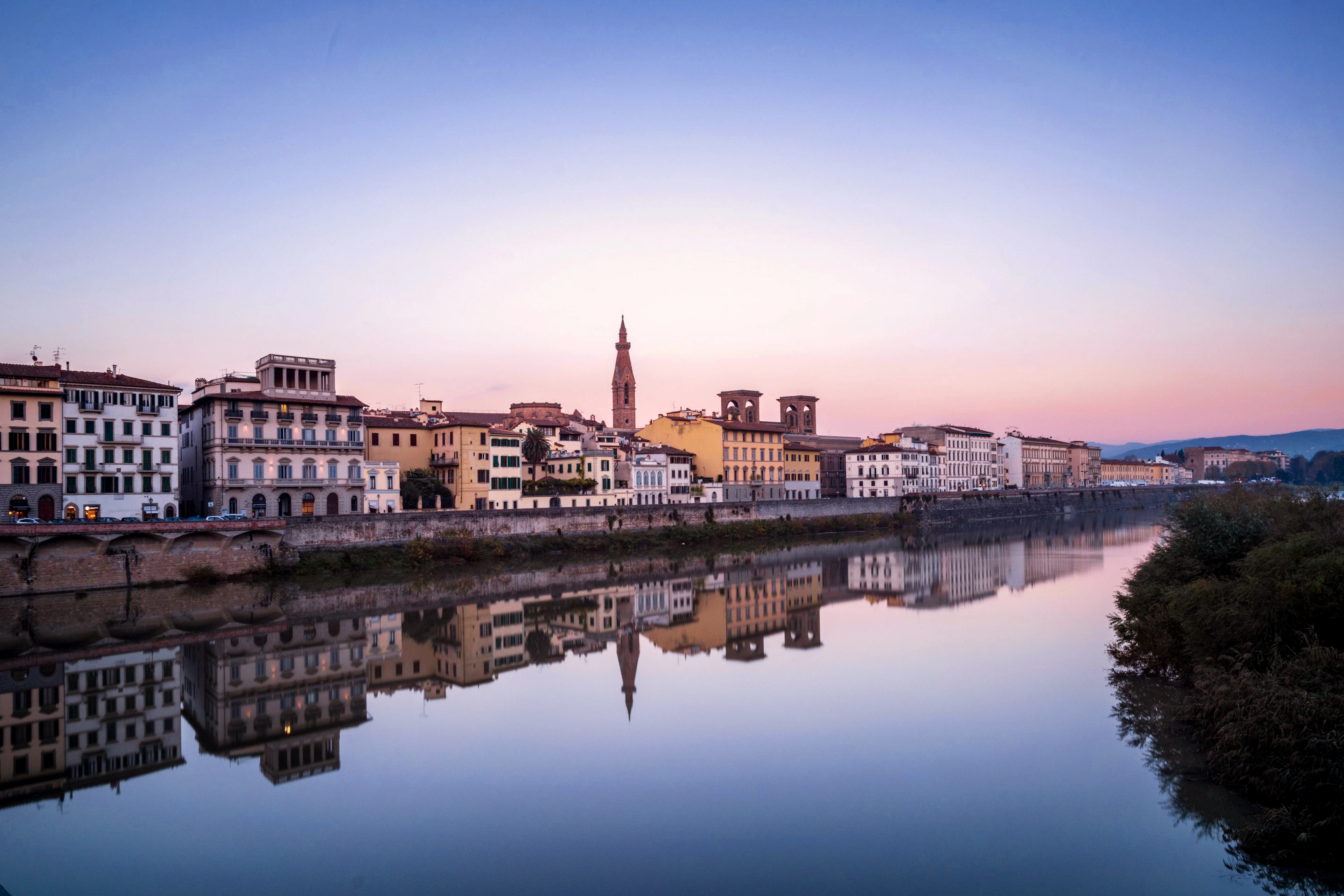 The Arno at dusk in Florence, a row of pale riverfront palazzi and the spire of Santa Croce mirrored in perfectly still water under a pink and lilac sky