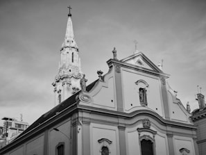A historic church with ornate architectural details and statues. The tall spire features a cross at the top, and the facade includes several decorative elements and sculptures. The image is captured in black and white.