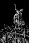 An intricately designed metallic sculpture of a person in historical clothing balances on a decorative structure with elaborate patterns and curves against a black background. The person appears to be in a dynamic pose with one arm raised, showcasing the detailed craftsmanship.