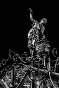 An intricately designed metallic sculpture of a person in historical clothing balances on a decorative structure with elaborate patterns and curves against a black background. The person appears to be in a dynamic pose with one arm raised, showcasing the detailed craftsmanship.