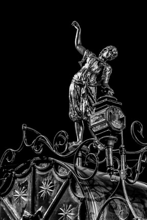 An intricately designed metallic sculpture of a person in historical clothing balances on a decorative structure with elaborate patterns and curves against a black background. The person appears to be in a dynamic pose with one arm raised, showcasing the detailed craftsmanship.