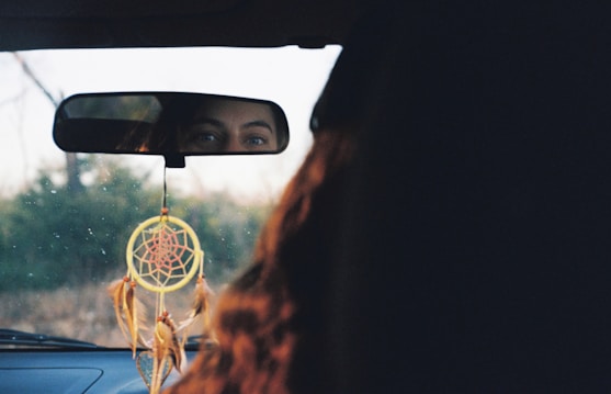 A rearview mirror captures the eyes of a person sitting in a car. A dreamcatcher with feathers and a circular web design hangs from the mirror. The background outside the car window is blurred, indicating a natural setting.
