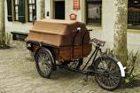 A vintage cargo tricycle with a brown covered storage compartment sits on a cobblestone street. The trike has a black frame and a brown leather seat. It is parked in front of a building with green window shutters and a door. Leafless plants border the sidewalk.