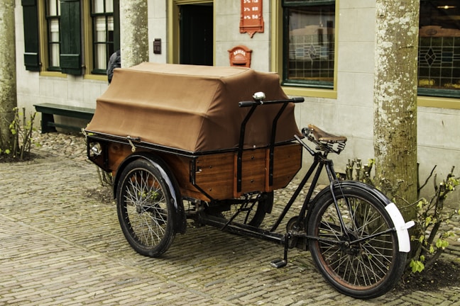 A vintage cargo tricycle with a brown covered storage compartment sits on a cobblestone street. The trike has a black frame and a brown leather seat. It is parked in front of a building with green window shutters and a door. Leafless plants border the sidewalk.
