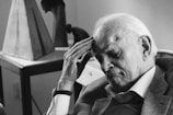 An elderly man is sitting thoughtfully, with his hand resting on his forehead. He appears deep in thought or contemplation, dressed in a suit jacket and shirt. The image is in black and white, with a moody and introspective atmosphere. The background is softly blurred, featuring abstract sculptures and a table.