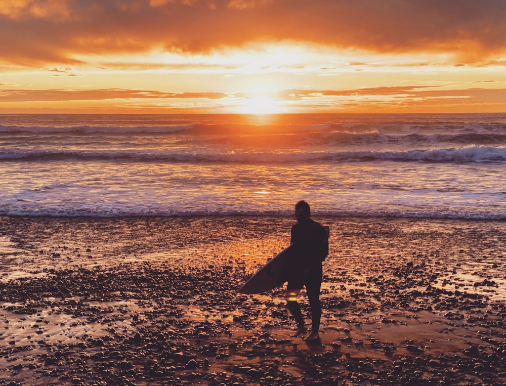 person holding surfing board walking towards sea during sunset