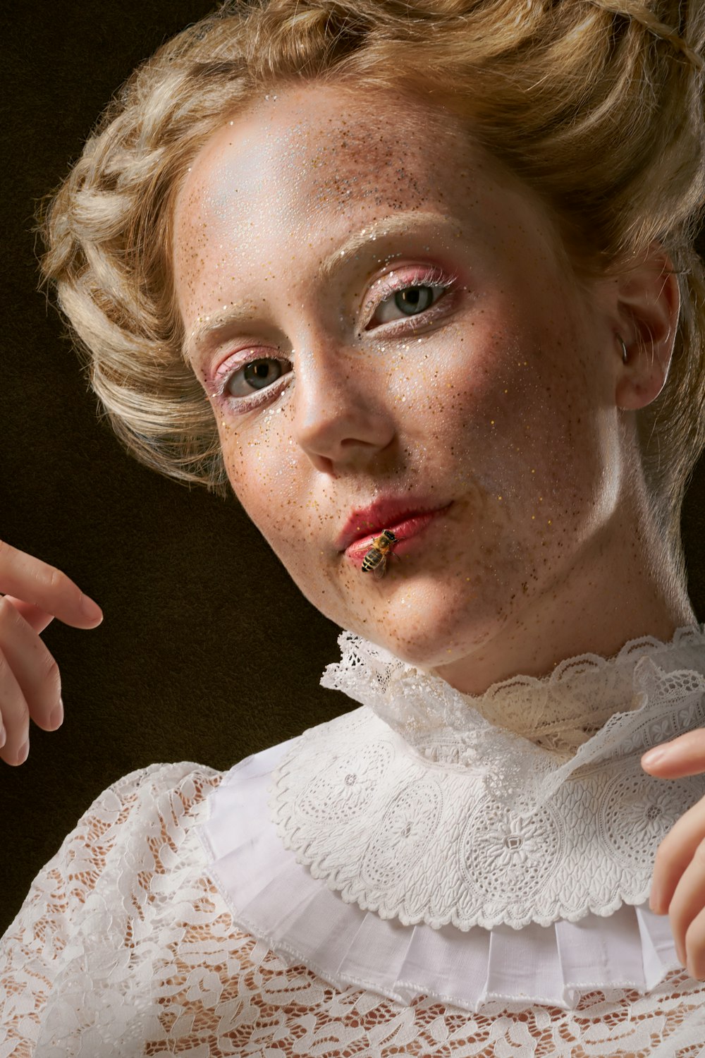 woman's portrait photo with white lace dress and glittered face having a bee on her lower lip