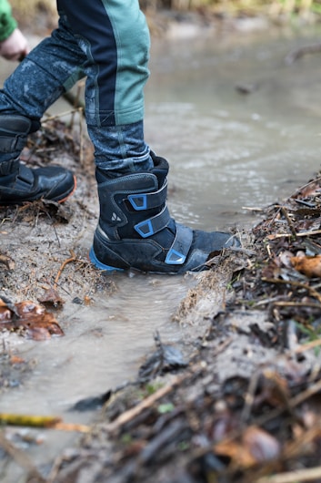 A person wearing sturdy blue and black boots steps through a shallow, muddy stream. The boots are wet and surrounded by fallen leaves and twigs on a dirt path. The water is slightly murky, with ripples around the foot creating a sense of movement.