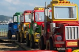 A row of colorful toy trucks is lined up on a wooden path by the sea. The trucks are primarily painted in yellow, red, green, and blue. The background features a blurry view of a beach and distant shoreline. One of the trucks has a visible sign labeled 'INTERCOOLER' and another labeled 'AULONA 01'.