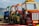 A row of colorful toy trucks is lined up on a wooden path by the sea. The trucks are primarily painted in yellow, red, green, and blue. The background features a blurry view of a beach and distant shoreline. One of the trucks has a visible sign labeled 'INTERCOOLER' and another labeled 'AULONA 01'.
