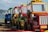 A row of colorful toy trucks is lined up on a wooden path by the sea. The trucks are primarily painted in yellow, red, green, and blue. The background features a blurry view of a beach and distant shoreline. One of the trucks has a visible sign labeled 'INTERCOOLER' and another labeled 'AULONA 01'.