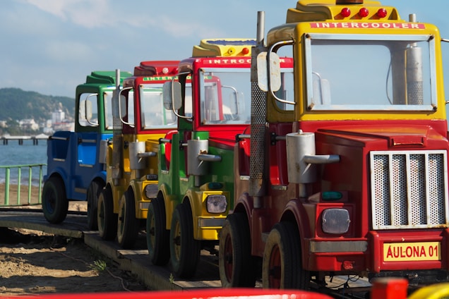 A row of colorful toy trucks is lined up on a wooden path by the sea. The trucks are primarily painted in yellow, red, green, and blue. The background features a blurry view of a beach and distant shoreline. One of the trucks has a visible sign labeled 'INTERCOOLER' and another labeled 'AULONA 01'.