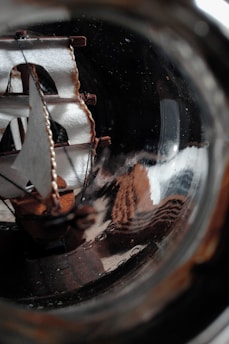 A miniature ship model with detailed sails is encased within a glass bottle, showcasing intricate craftsmanship. The texture of the sails and the rigging creates a dynamic contrast with the smooth, reflective surface of the glass, giving an impression of motion and artistry.