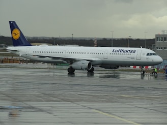 An airplane from Lufthansa is stationary on an airport runway. The aircraft is mostly white with a dark blue tail featuring the airline's logo. The tarmac appears wet, indicating recent rainfall, and the sky is overcast with gray clouds.