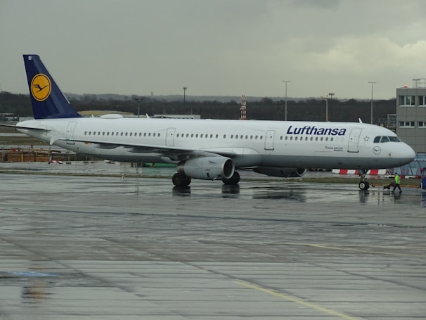 An airplane from Lufthansa is stationary on an airport runway. The aircraft is mostly white with a dark blue tail featuring the airline's logo. The tarmac appears wet, indicating recent rainfall, and the sky is overcast with gray clouds.