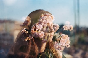 A double exposure photograph blending a woman's face with pink cherry blossoms. The soft focus and overlapping elements create a dreamlike effect. The background appears blurred, enhancing the surreal quality.