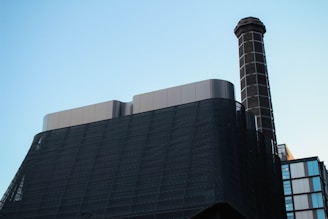 A modern industrial building features a tall chimney with a checkered pattern extending upwards. The structure is constructed with dark, sleek materials and smooth metallic surfaces. The background is a clear sky, lending a minimalist contrast to the industrial elements.