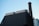 A modern industrial building features a tall chimney with a checkered pattern extending upwards. The structure is constructed with dark, sleek materials and smooth metallic surfaces. The background is a clear sky, lending a minimalist contrast to the industrial elements.
