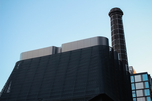 A modern industrial building features a tall chimney with a checkered pattern extending upwards. The structure is constructed with dark, sleek materials and smooth metallic surfaces. The background is a clear sky, lending a minimalist contrast to the industrial elements.
