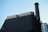 A modern industrial building features a tall chimney with a checkered pattern extending upwards. The structure is constructed with dark, sleek materials and smooth metallic surfaces. The background is a clear sky, lending a minimalist contrast to the industrial elements.