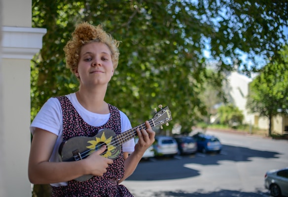 A person with curly hair holds a decorated ukulele while standing outdoors, surrounded by lush green trees. The background includes parked cars and a blurred street scene, creating a relaxed and casual atmosphere.