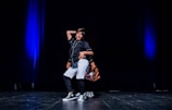 A young person is performing a hip-hop dance on stage, wearing casual dance attire and sneakers. The stage is dimly lit with blue spotlighting, giving a dramatic effect to the performance.