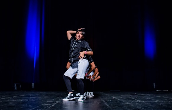 A young person is performing a hip-hop dance on stage, wearing casual dance attire and sneakers. The stage is dimly lit with blue spotlighting, giving a dramatic effect to the performance.