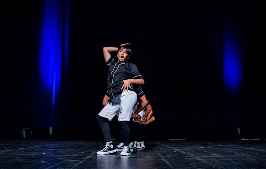 A young person is performing a hip-hop dance on stage, wearing casual dance attire and sneakers. The stage is dimly lit with blue spotlighting, giving a dramatic effect to the performance.