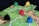 A board game setup with colorful wooden game pieces, known as meeples, placed on square tiles. The tiles depict green fields and small sections of villages or roads.