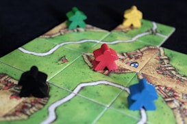 A board game setup with colorful wooden game pieces, known as meeples, placed on square tiles. The tiles depict green fields and small sections of villages or roads.