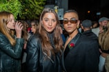 A group of stylishly dressed people are gathered at an outdoor night event. In the foreground, two individuals are posing for the camera, one wearing a black jacket with a rose accessory and sunglasses, while the other has long hair and a leather jacket. The background shows more people, some holding drinks, under a tree with green leaves.