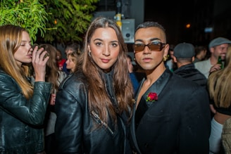 A group of stylishly dressed people are gathered at an outdoor night event. In the foreground, two individuals are posing for the camera, one wearing a black jacket with a rose accessory and sunglasses, while the other has long hair and a leather jacket. The background shows more people, some holding drinks, under a tree with green leaves.