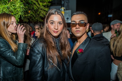 A group of stylishly dressed people are gathered at an outdoor night event. In the foreground, two individuals are posing for the camera, one wearing a black jacket with a rose accessory and sunglasses, while the other has long hair and a leather jacket. The background shows more people, some holding drinks, under a tree with green leaves.